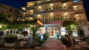 A multi-story hotel with a restaurant on the ground floor, outdoor seating, and a well-lit entrance. - Hotel Maritimo (Makarska)