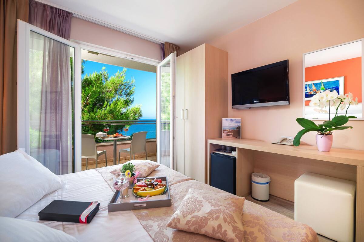 A hotel room with a bed, a desk, a TV, a balcony with a view of the sea, and a dining table with food.