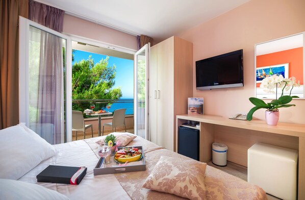 Una habitación de hotel con cama, escritorio, televisor, balcón con vista al mar y una mesa de comedor con comida.