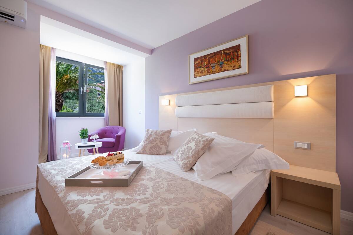A bedroom with a bed, a nightstand, a window with curtains, a small table with a plant, and a painting on the wall.