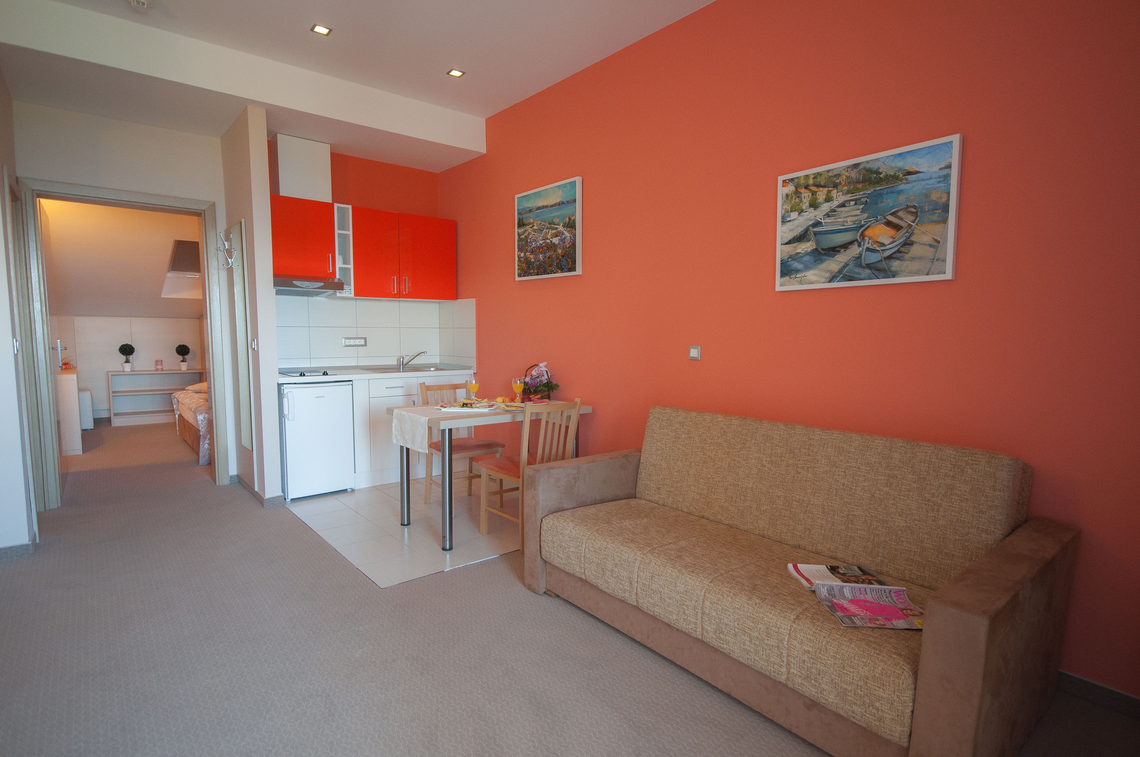 A modern living room with a beige sofa, a dining area with a table and chairs, and a kitchen with white cabinets and a red accent wall.