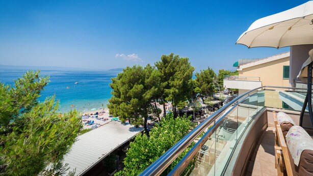 A balcony with a glass railing overlooking a beach with umbrellas and people. - Hotel Maritimo (Makarska)