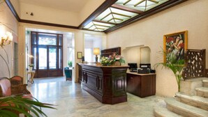 A spacious lobby with a marble floor, a reception desk, a staircase, and a large window with a view of the outdoors. - Grand Hotel Regina (Salsomaggiore Terme)