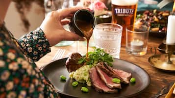 A person pouring sauce over a plate of food, including meat and greens, with a beer glass and candle in the background.