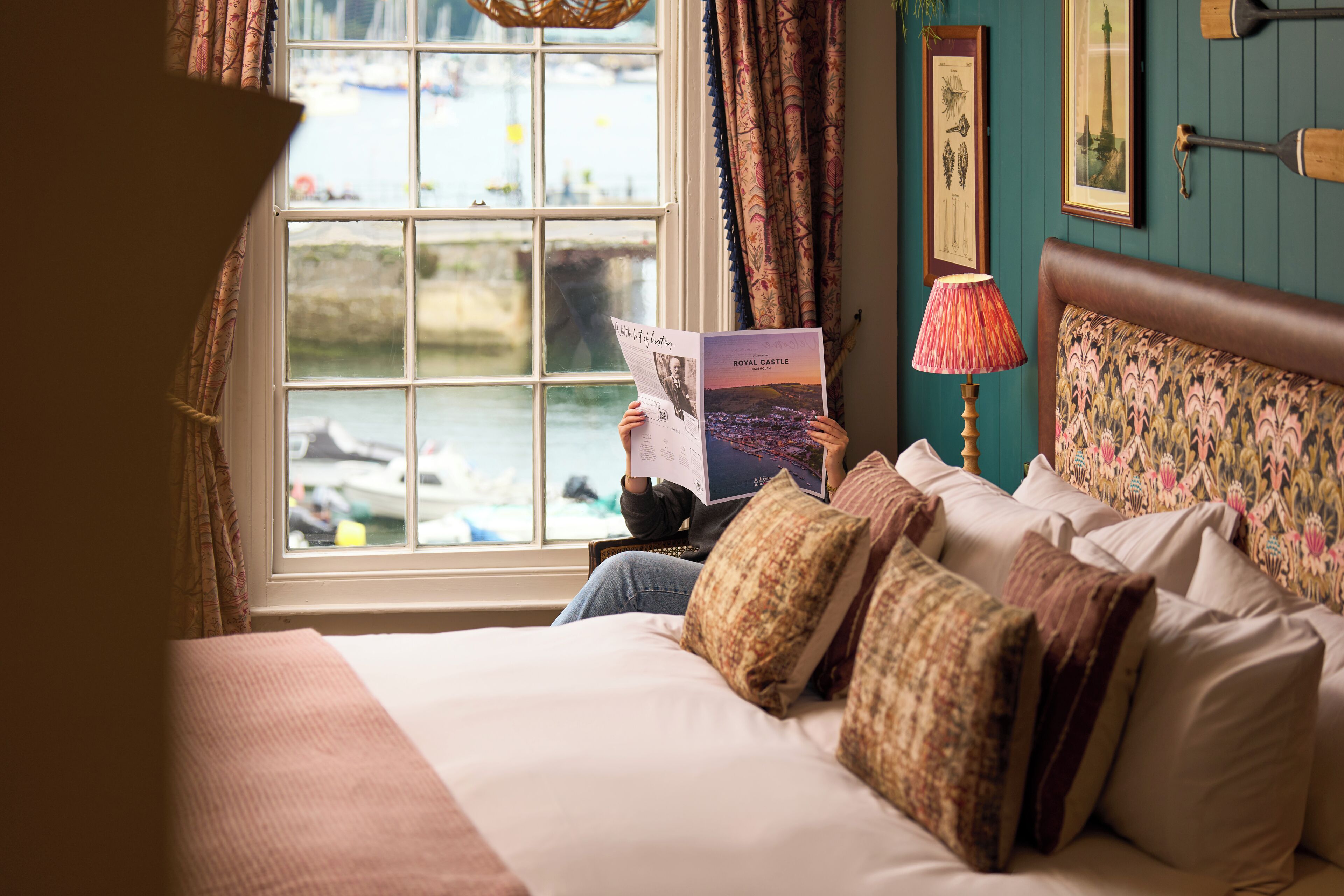 A bed with a view of boats through a window, a lamp, and a framed picture on the wall.