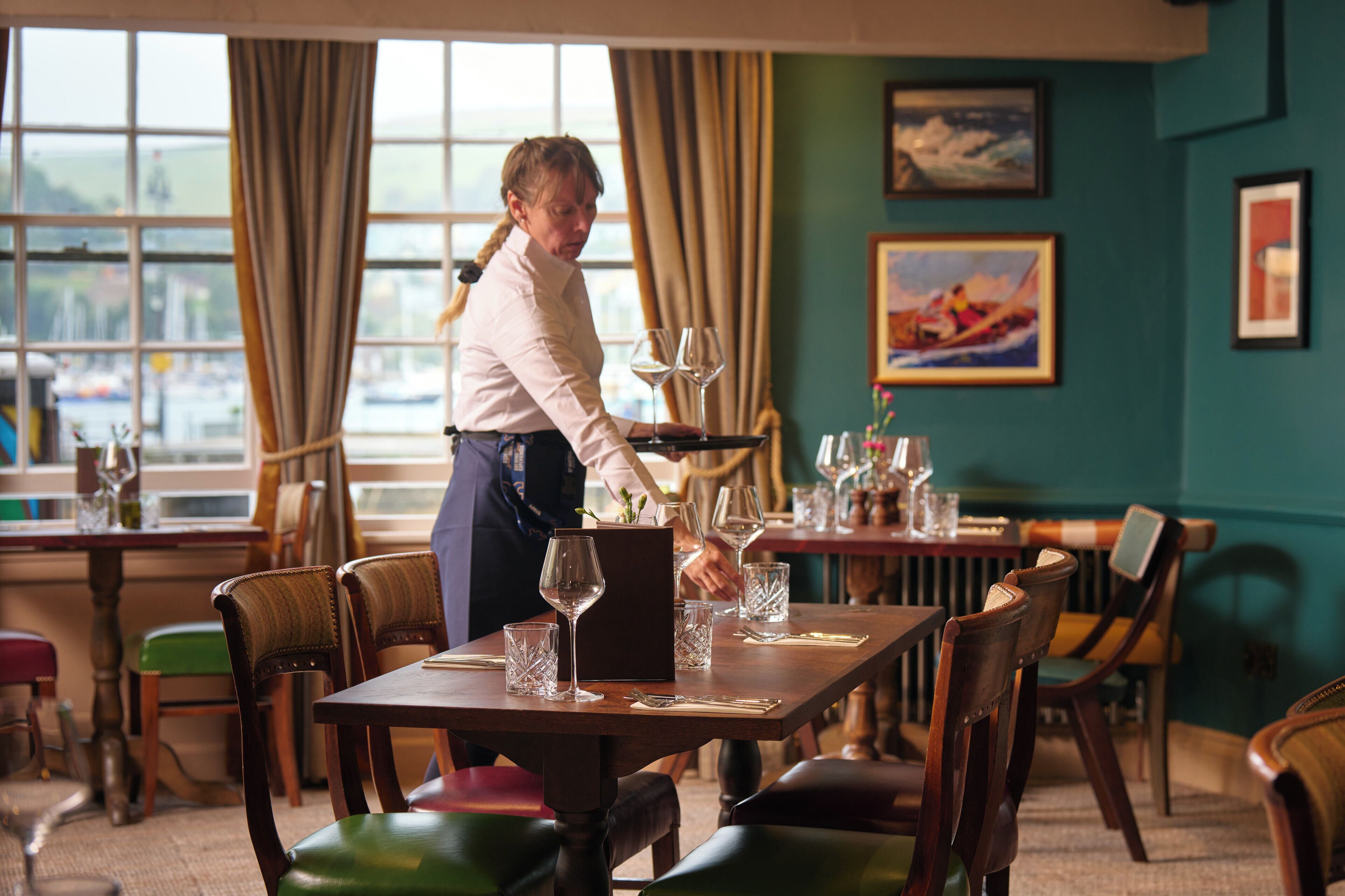 Een restaurant met houten tafels en stoelen, een serveerster die een tafel bedient, en uitzicht op de zee door grote ramen.