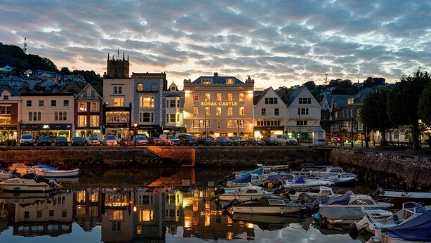 A waterfront town with historic buildings, including a prominent hotel named "Royal Castle." Boats are docked along the water's edge, and the area is reflected in the calm water. - Royal Castle Hotel (Dartmouth)