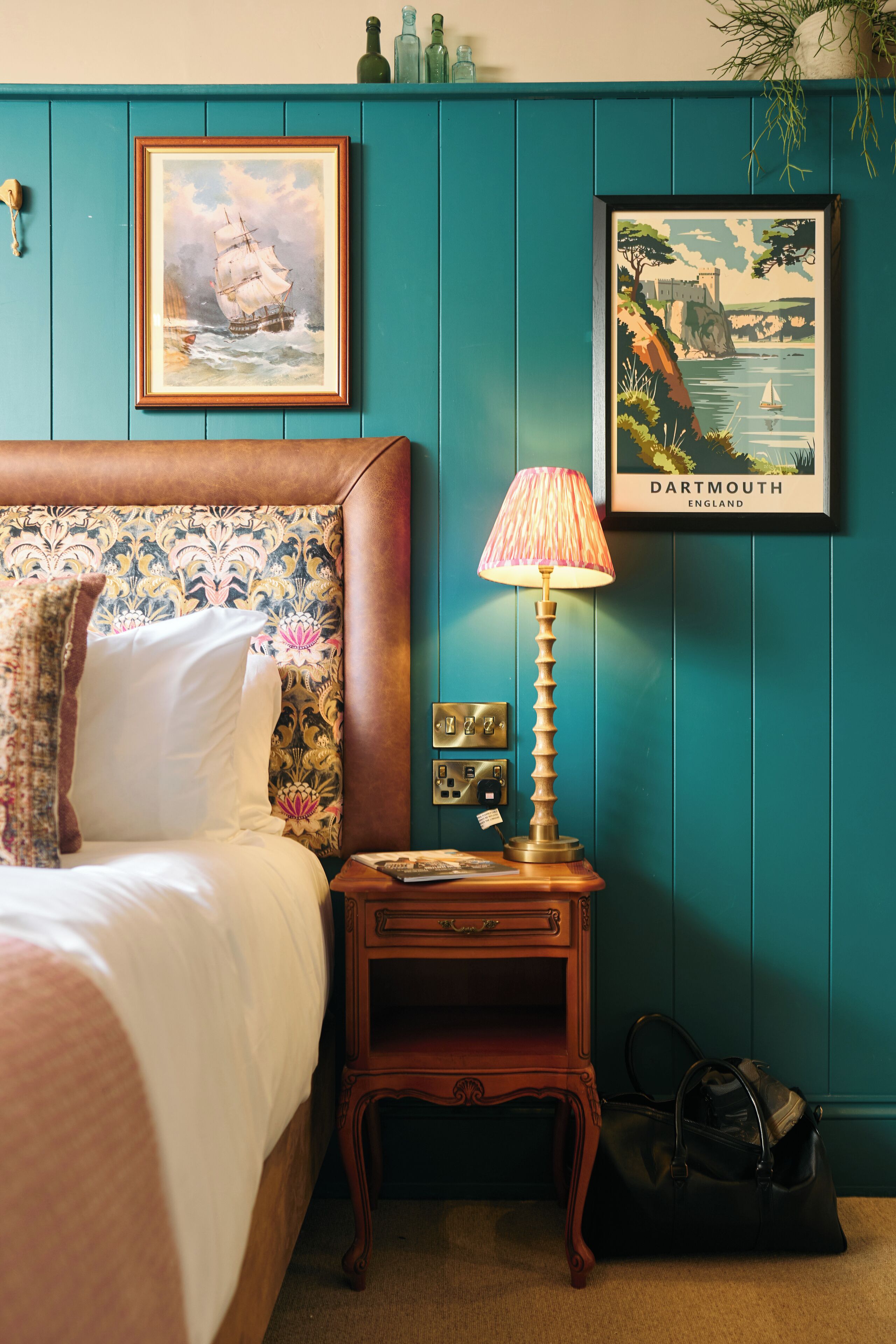 A bedroom with a bed, a nightstand with a lamp, a framed picture of a sailing ship, and a framed picture of a coastal scene.
