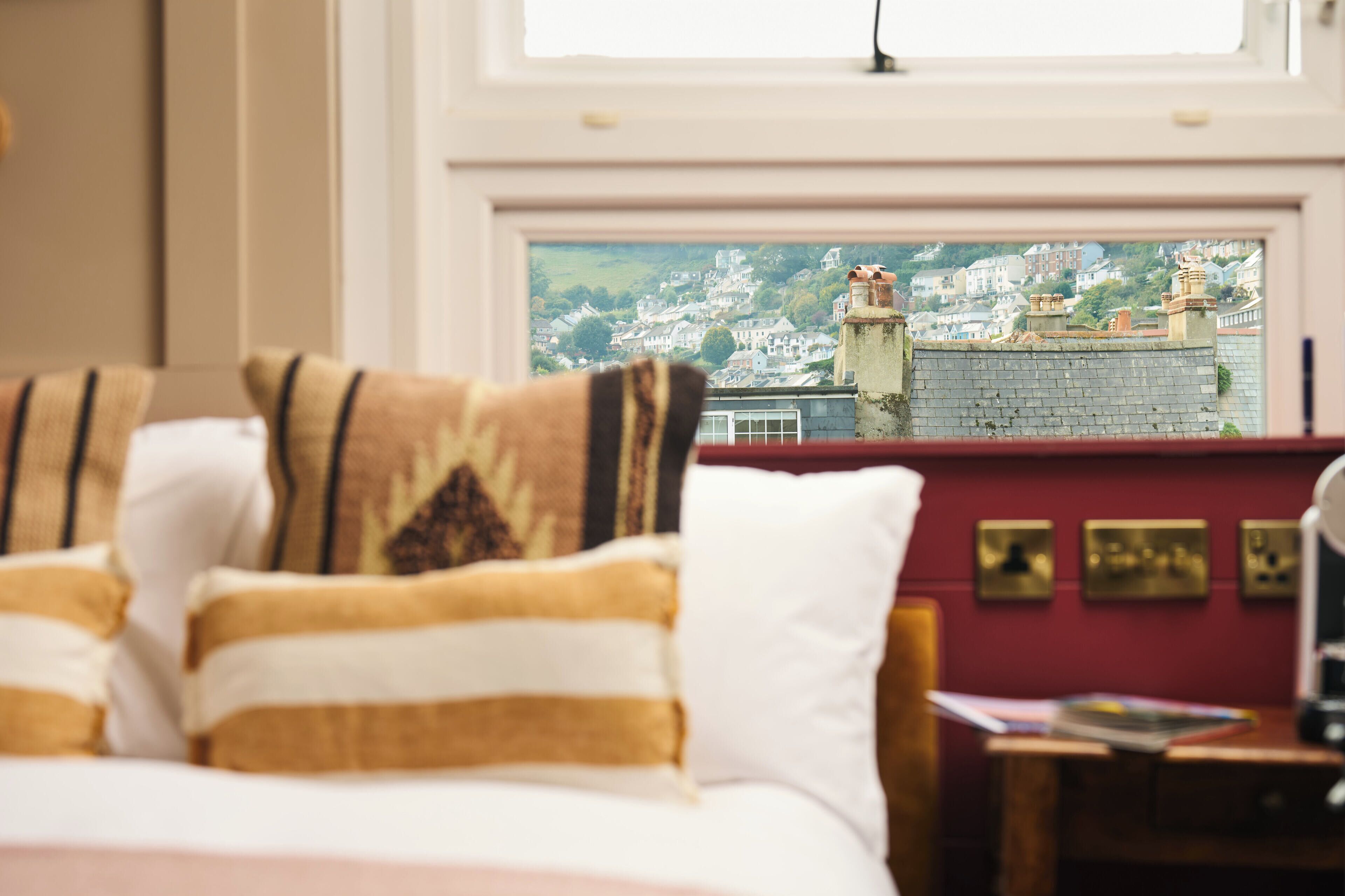 A hotel room with a bed, pillows, a desk, and a view of a town through a window.
