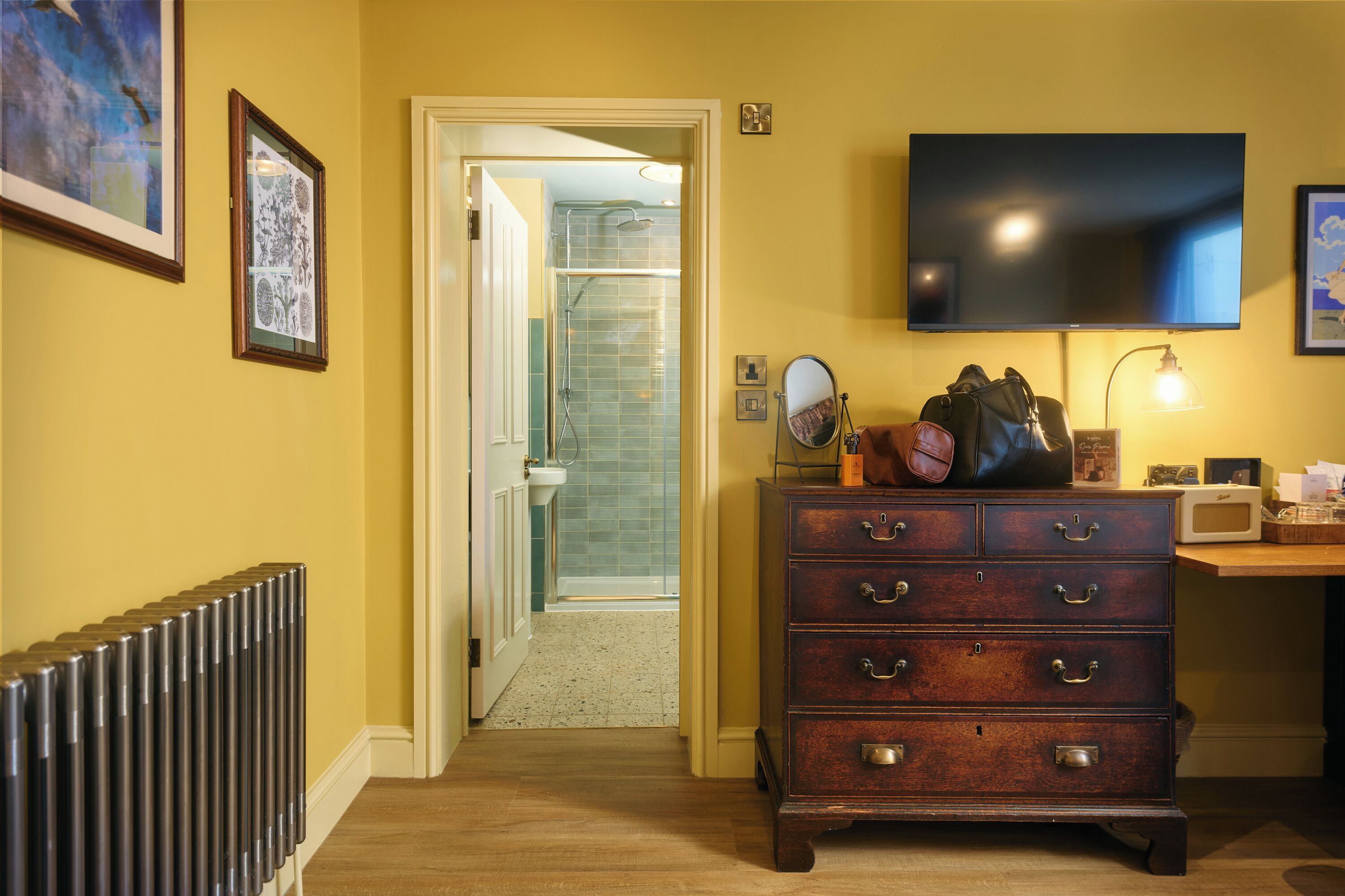 A hotel room with a wooden dresser, a television mounted on the wall, a radiator, and a framed picture on the wall.