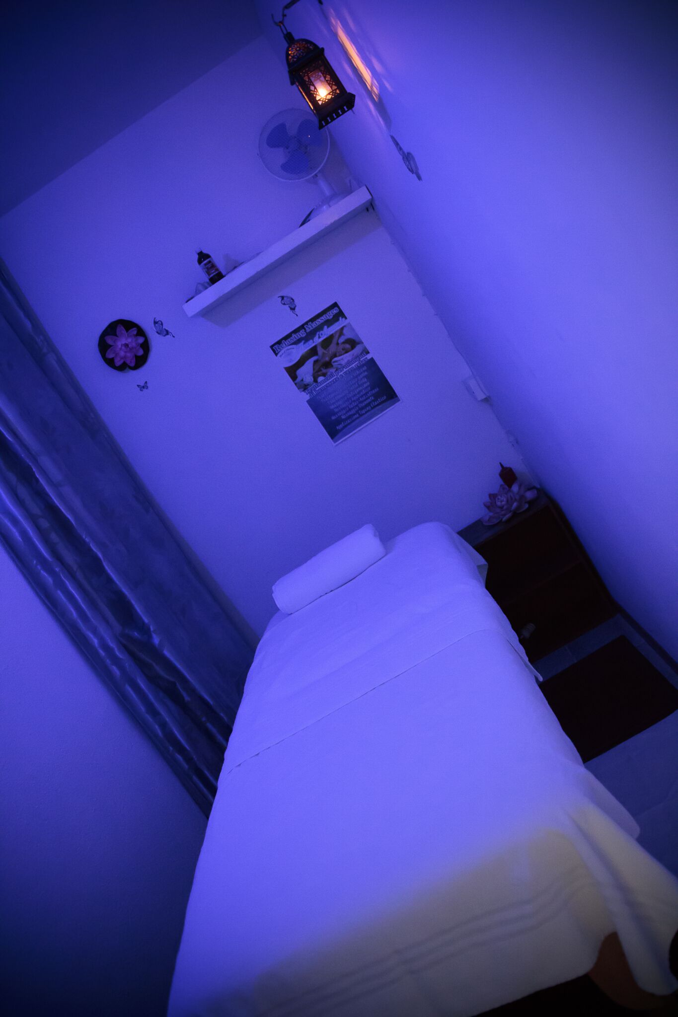 A room with a bed, a wall-mounted shelf with items, a poster, and a small fan.