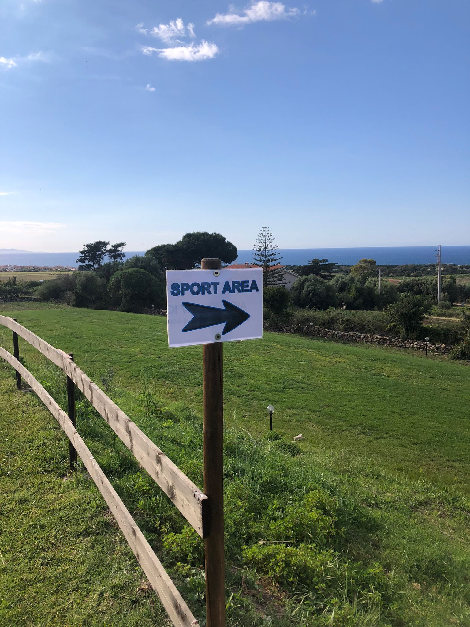 A sign indicating a sport area with a directional arrow, set against a backdrop of a grassy field, a wooden fence, and a clear sky.