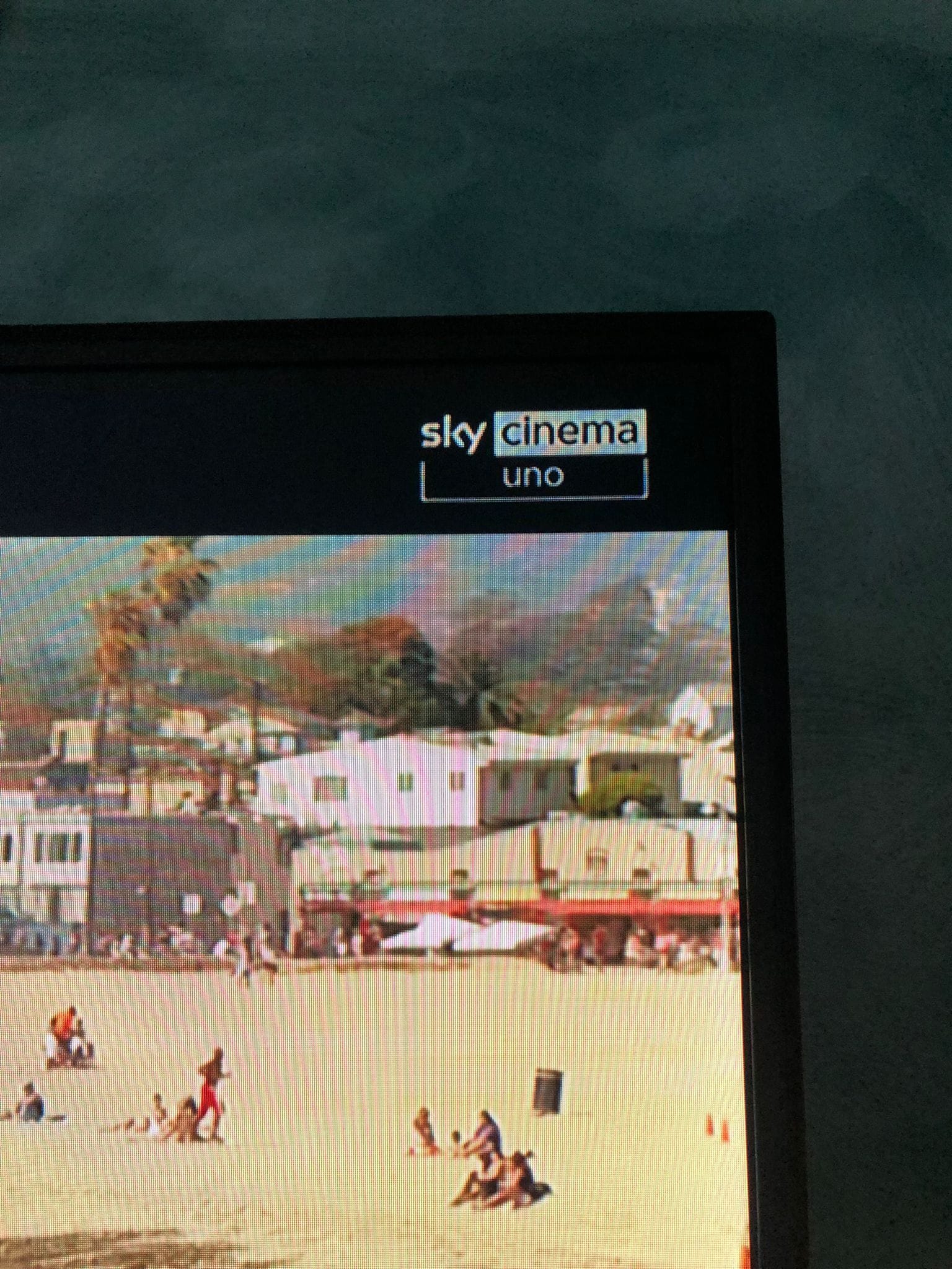 Een televisiescherm toont een beeld met gebouwen en mensen, met een 'Sky Cinema Uno'-logo aan de bovenkant.