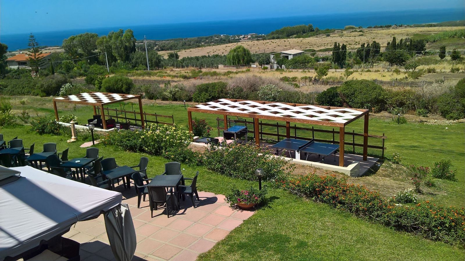 An outdoor seating area with tables and chairs, a ping pong table, and a view of the sea and hills.