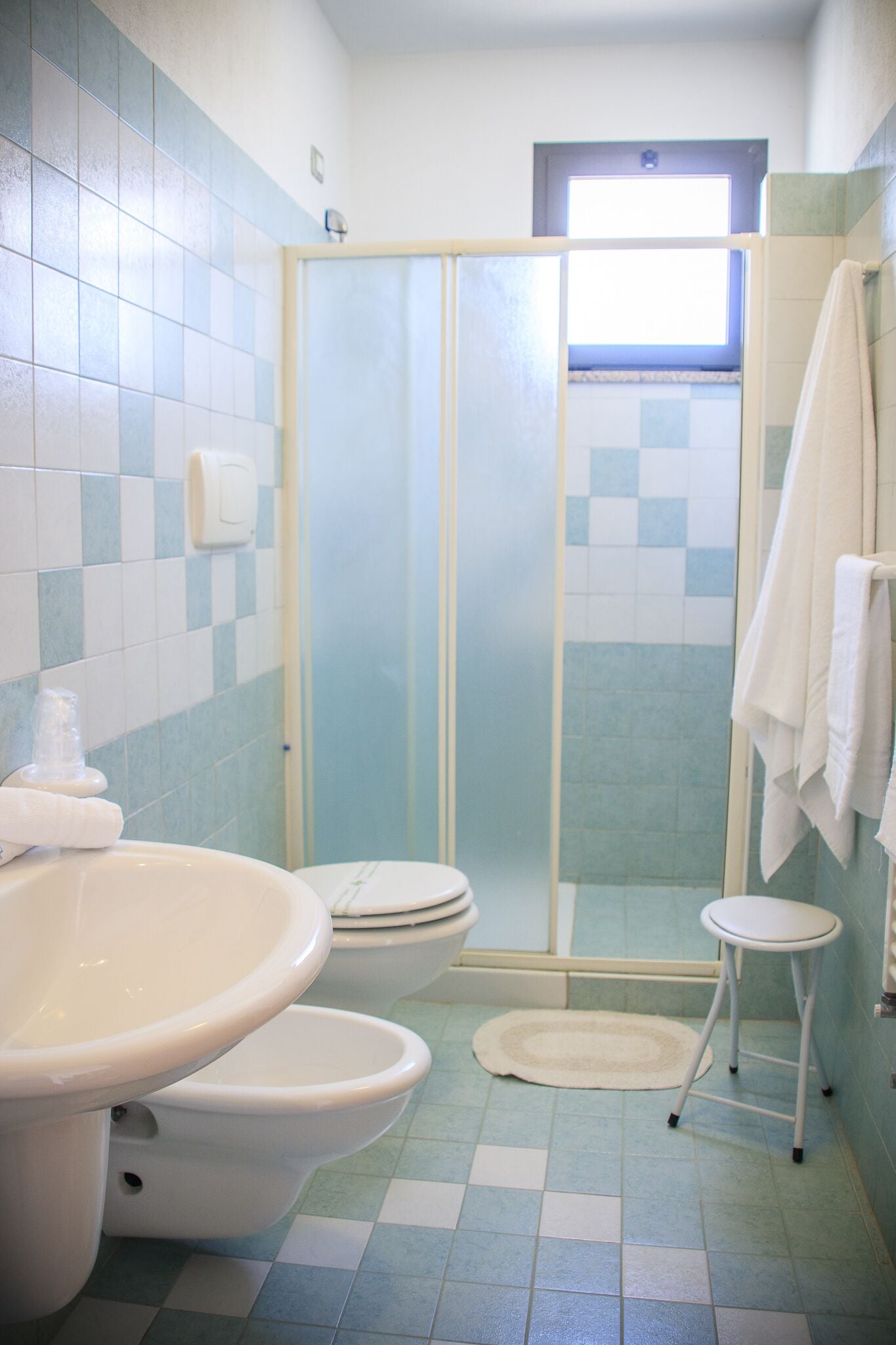 A bathroom with a shower enclosure, a sink, two toilets, and a small stool.