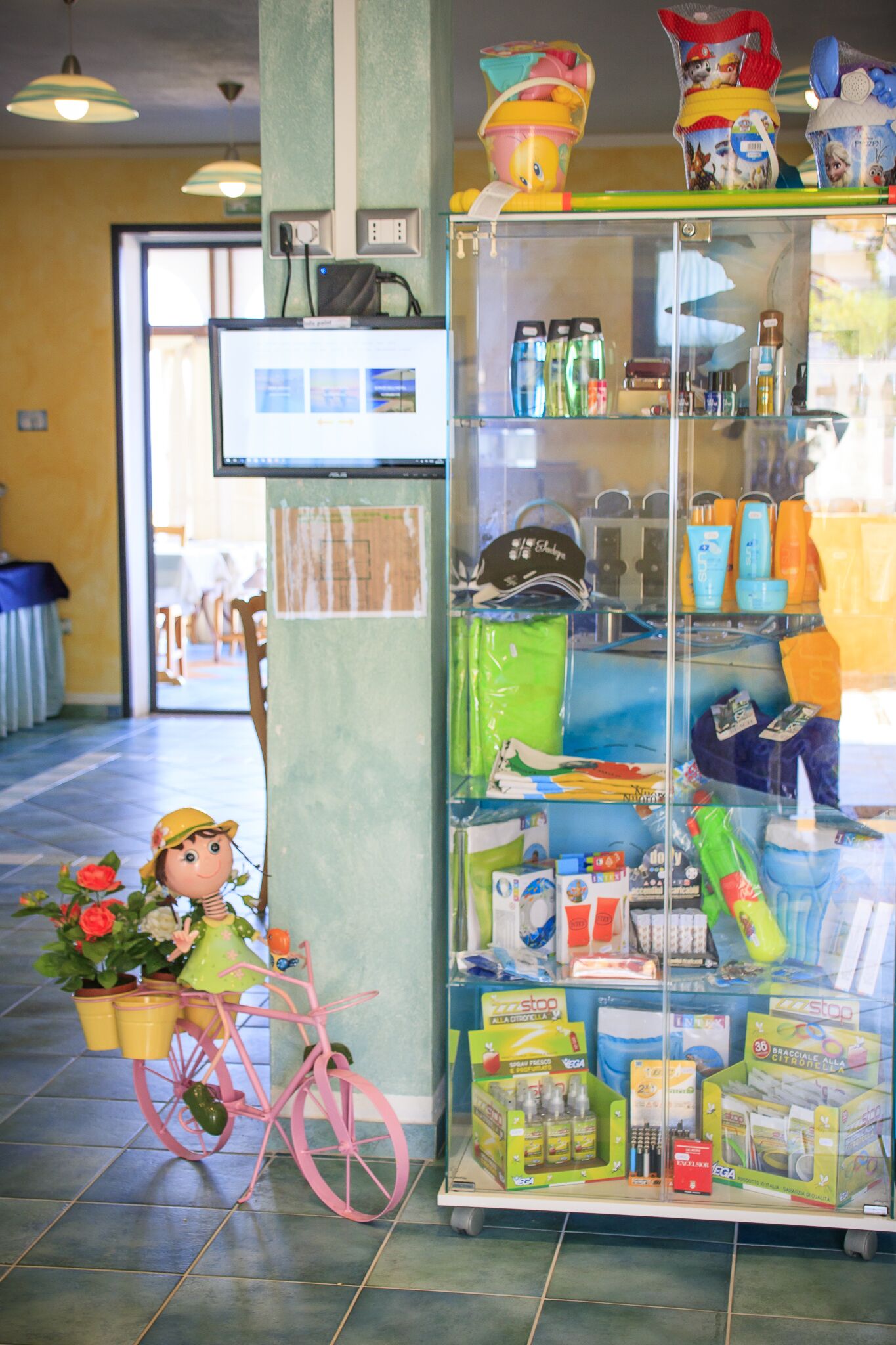 A store with shelves stocked with various products, a pink bicycle, and a computer monitor on a stand.