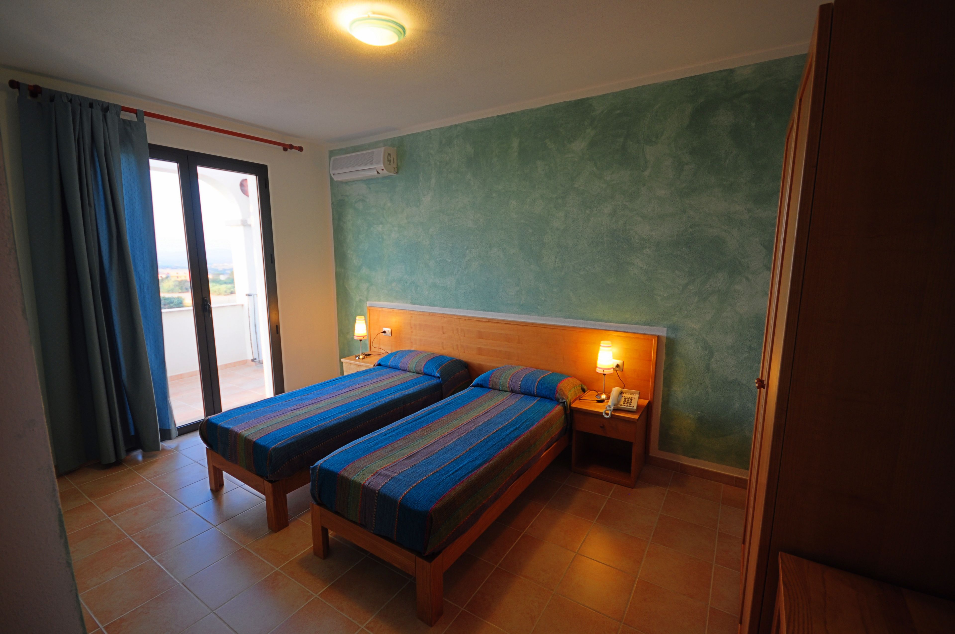 A room with two beds, a nightstand with a lamp, and a view through a sliding door to an outdoor area.