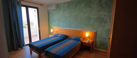 A room with two beds, a nightstand with a lamp, and a view through a sliding door to an outdoor area.