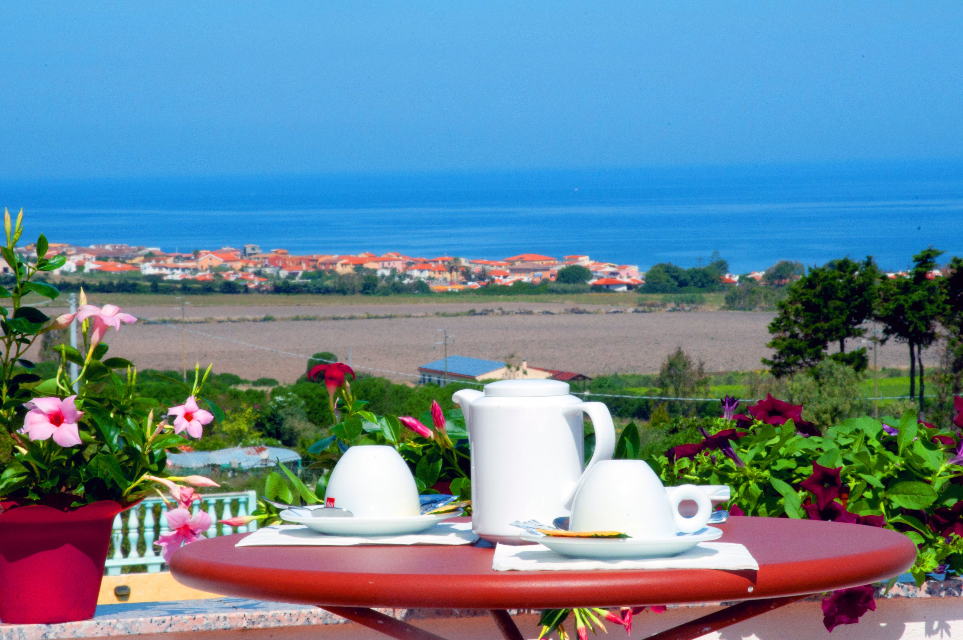 Ein Tisch mit weißen Tassen und einer weißen Kanne, vor einer malerischen Aussicht auf das Meer und eine Küstenstadt.