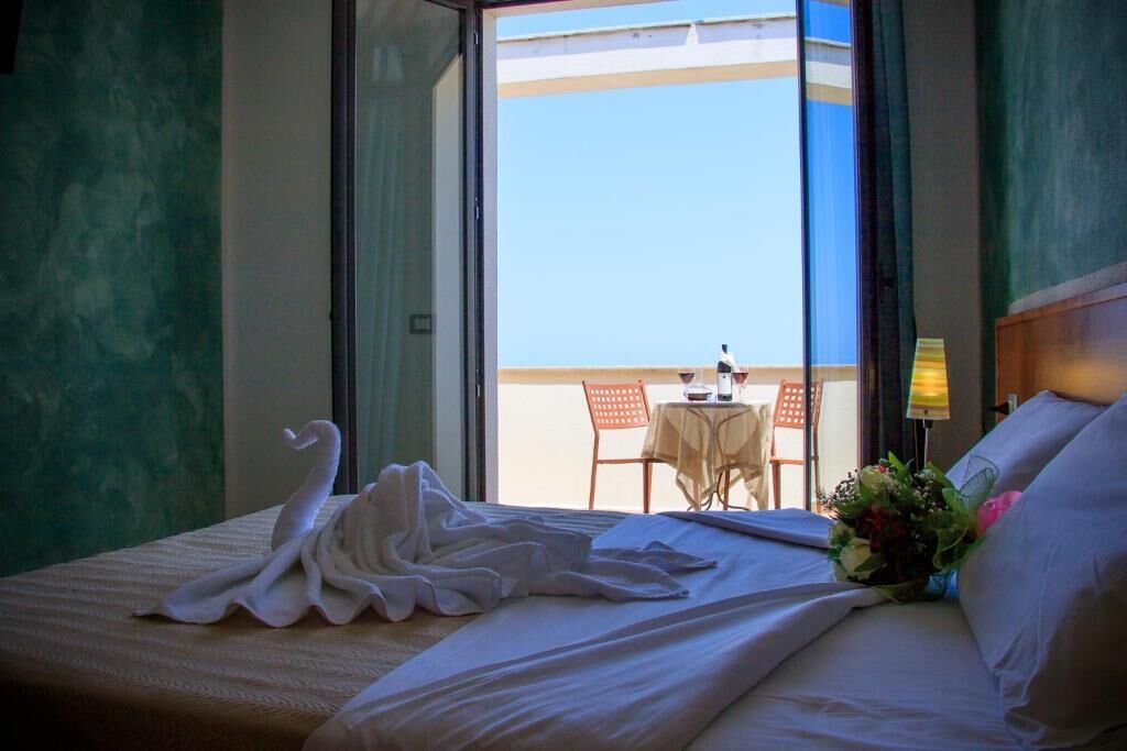 Una camera d'albergo con un letto, una finestra vista spiaggia, un tavolo e una sedia, e un vaso con fiori.