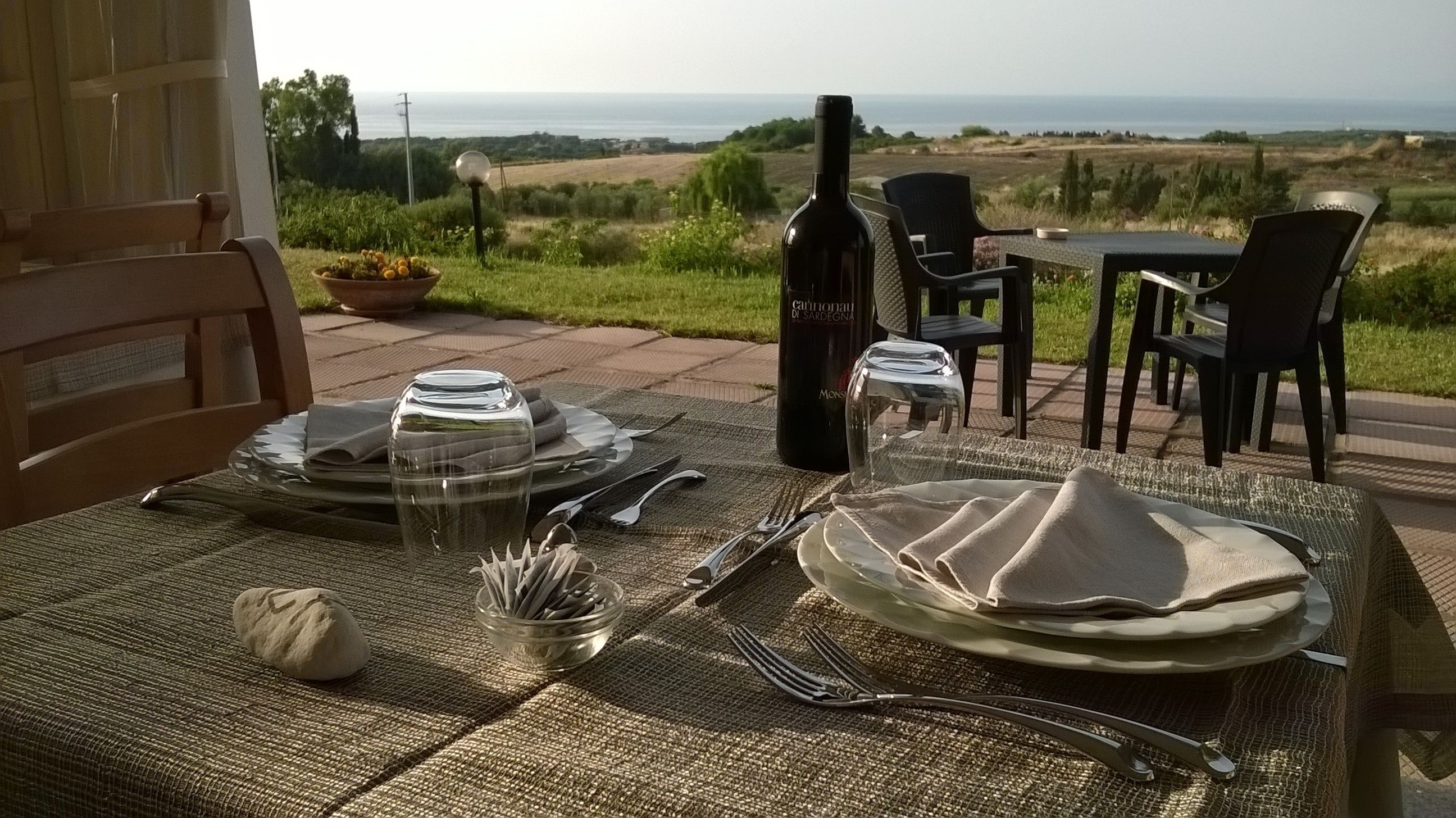 An outdoor dining setup with a view of the sea, a bottle of wine, and a table set with plates, cutlery, and a bowl of bread.