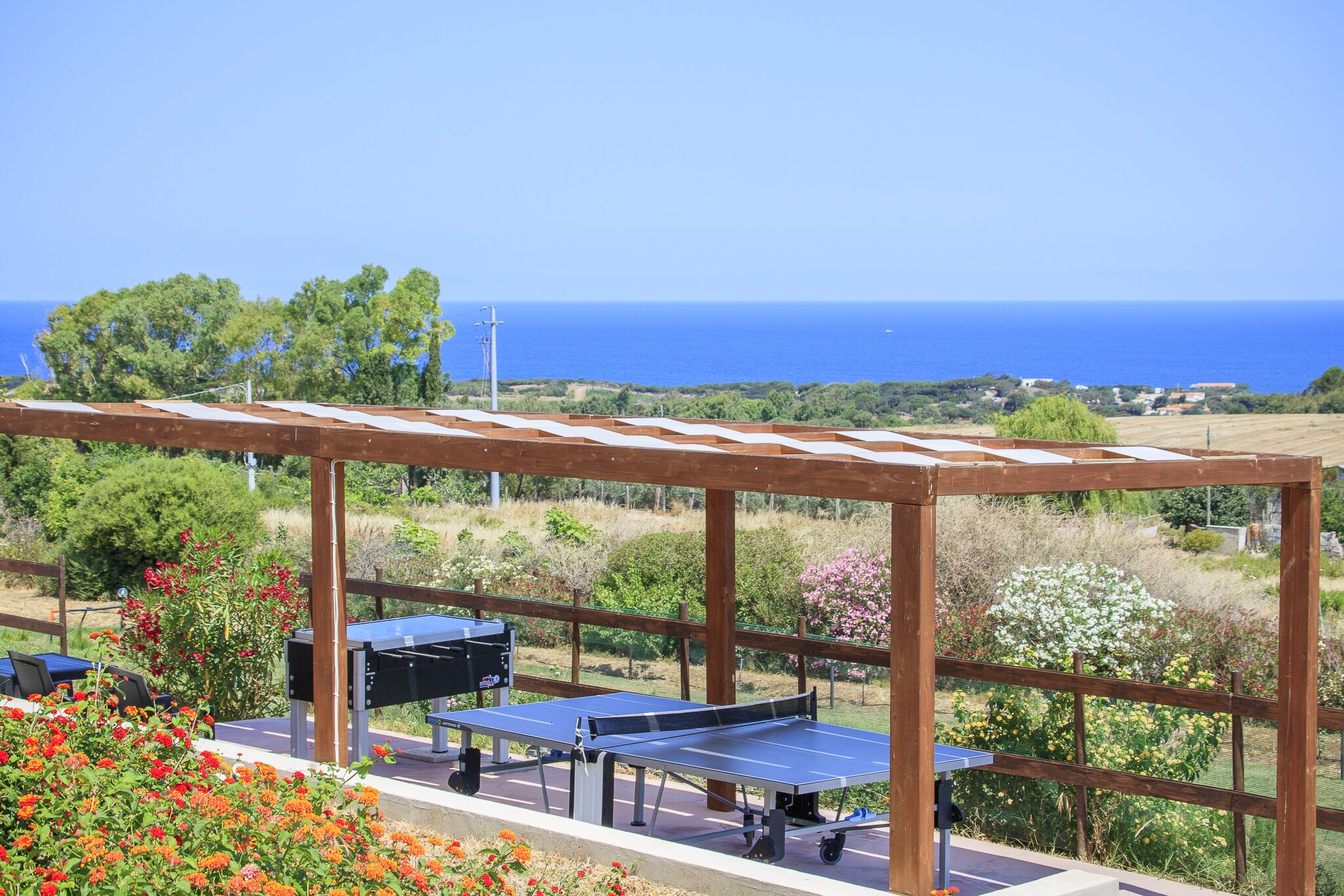 An outdoor ping pong table set up under a wooden pergola with a view of the sea.