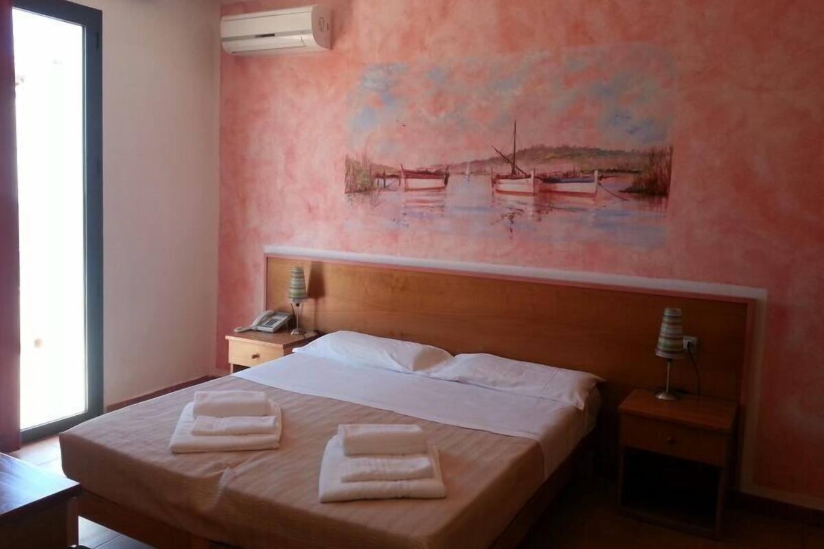 A hotel room with a bed, a nightstand, an air conditioning unit, and a painting on the wall.
