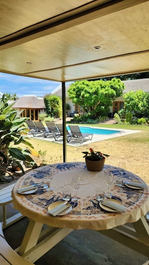 A covered outdoor dining area with a table set for four, overlooking a swimming pool and a garden. - KaapsePracht (Cape Town)
