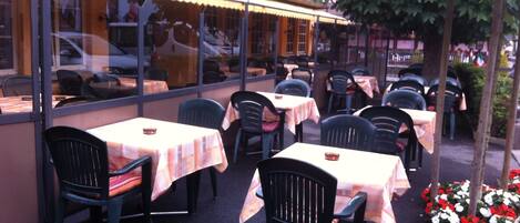 An outdoor cafe with tables and chairs, a building with a window, and a tree.