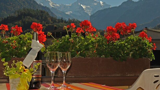 A table with a bottle and glasses, red flowers, and a mountainous backdrop. - Hotel Post Hardermannli (Unterseen)
