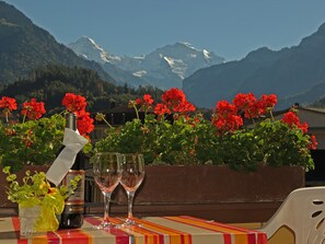 A table with a bottle and glasses, red flowers, and a mountainous backdrop. - Hotel Post Hardermannli (Unterseen)