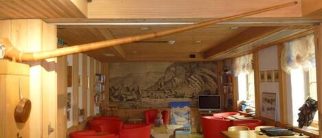 A cozy room with wooden walls, a large mural, red seating, a television, and a wooden table.