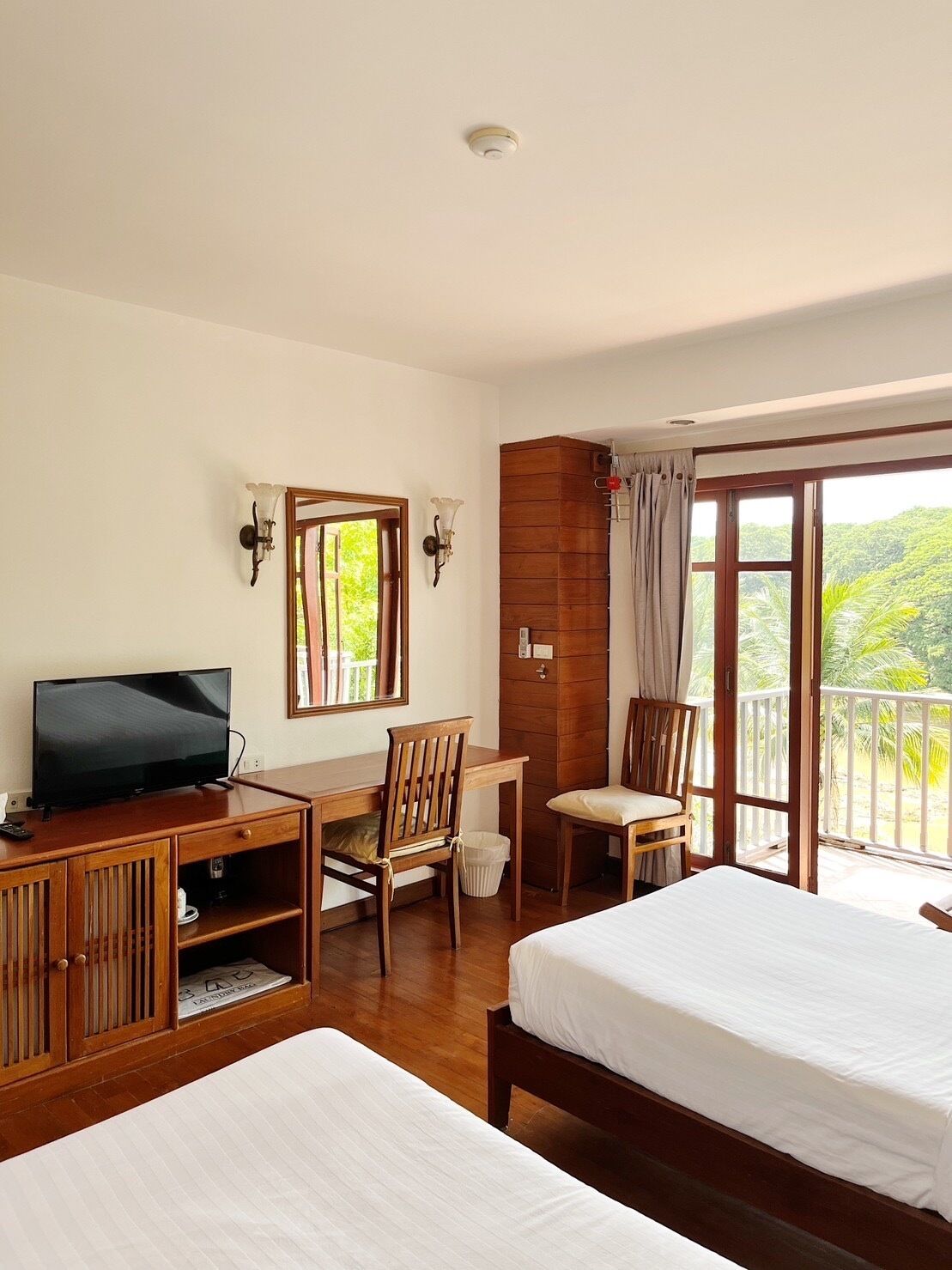 A hotel room with two beds, a TV, a desk, and a balcony with a view of greenery.
