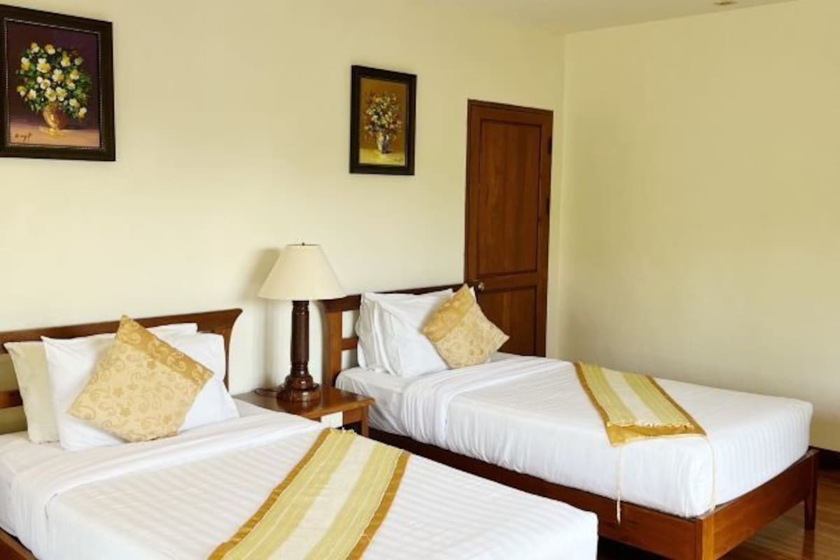 Una habitación de hotel con dos camas individuales, un cabecero de madera, una mesita de noche con una lámpara y dos cuadros enmarcados con motivos florales en la pared.