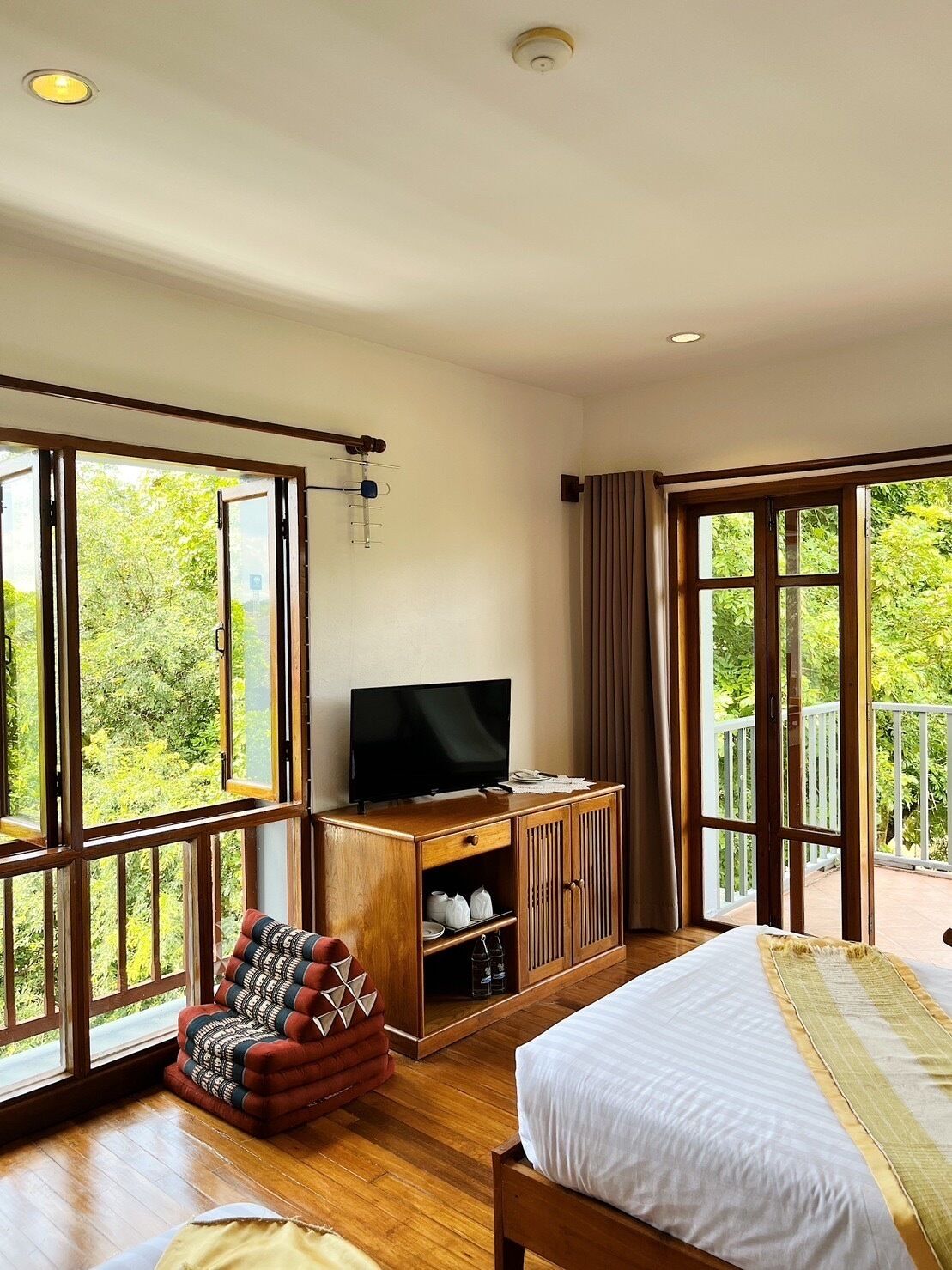 A bedroom with a bed, a TV on a wooden cabinet, a chair, and large windows with a balcony.