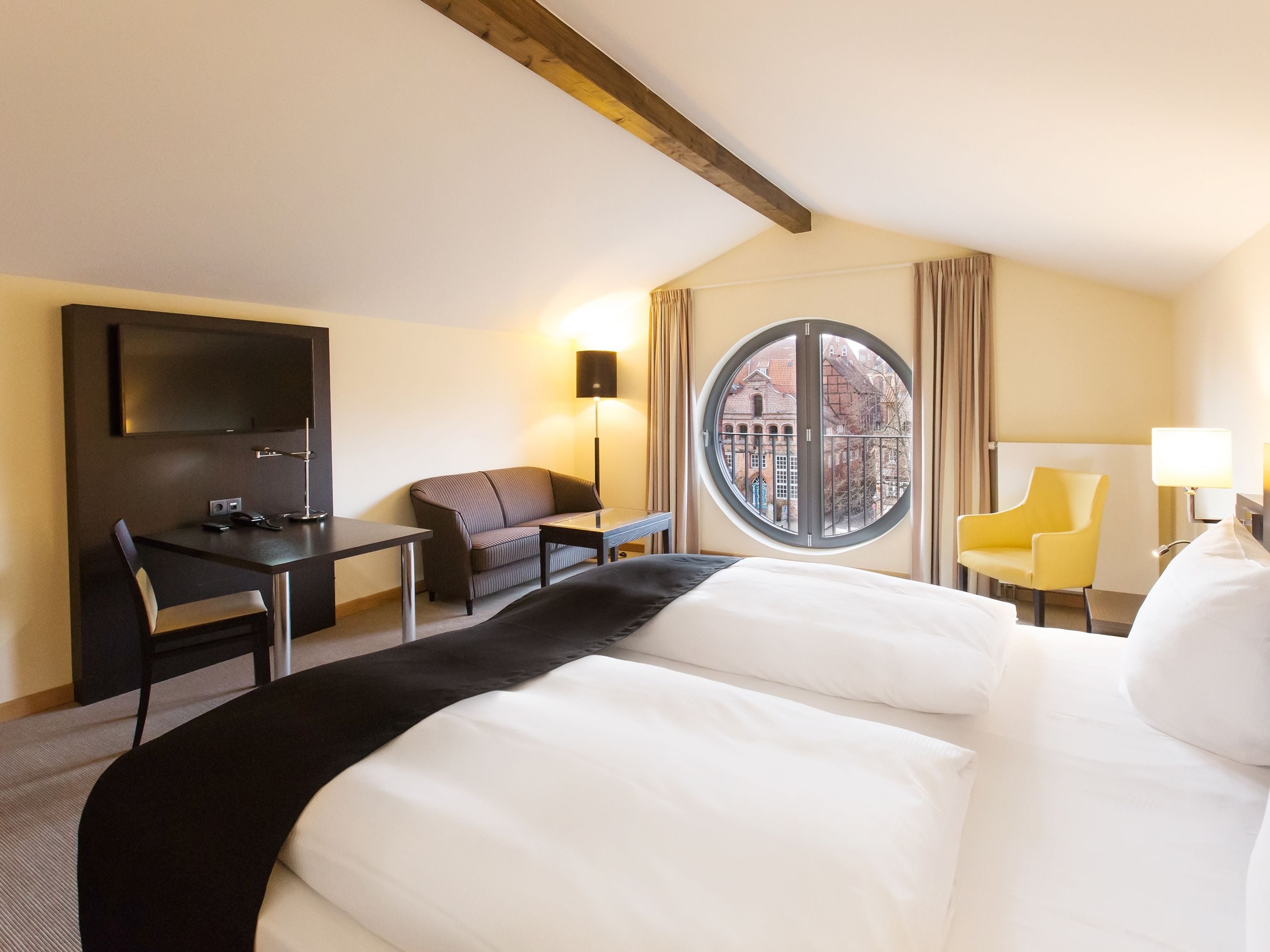 Quarto de hotel com cama, escrivaninha, cadeira, televisão e janela com vista para edifícios.