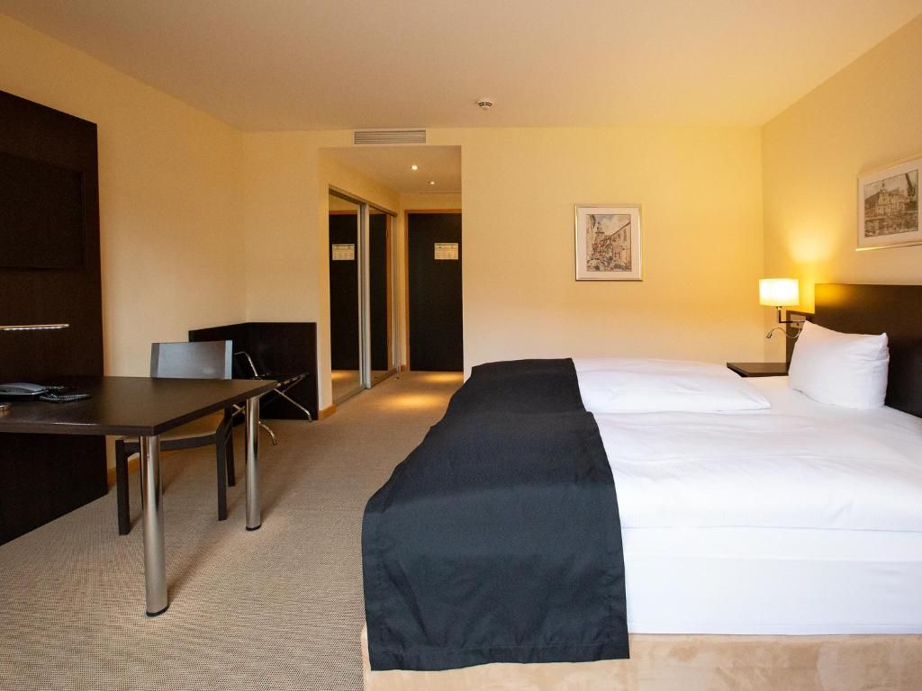 A hotel room with a bed, a desk, a chair, and a nightstand with a lamp.