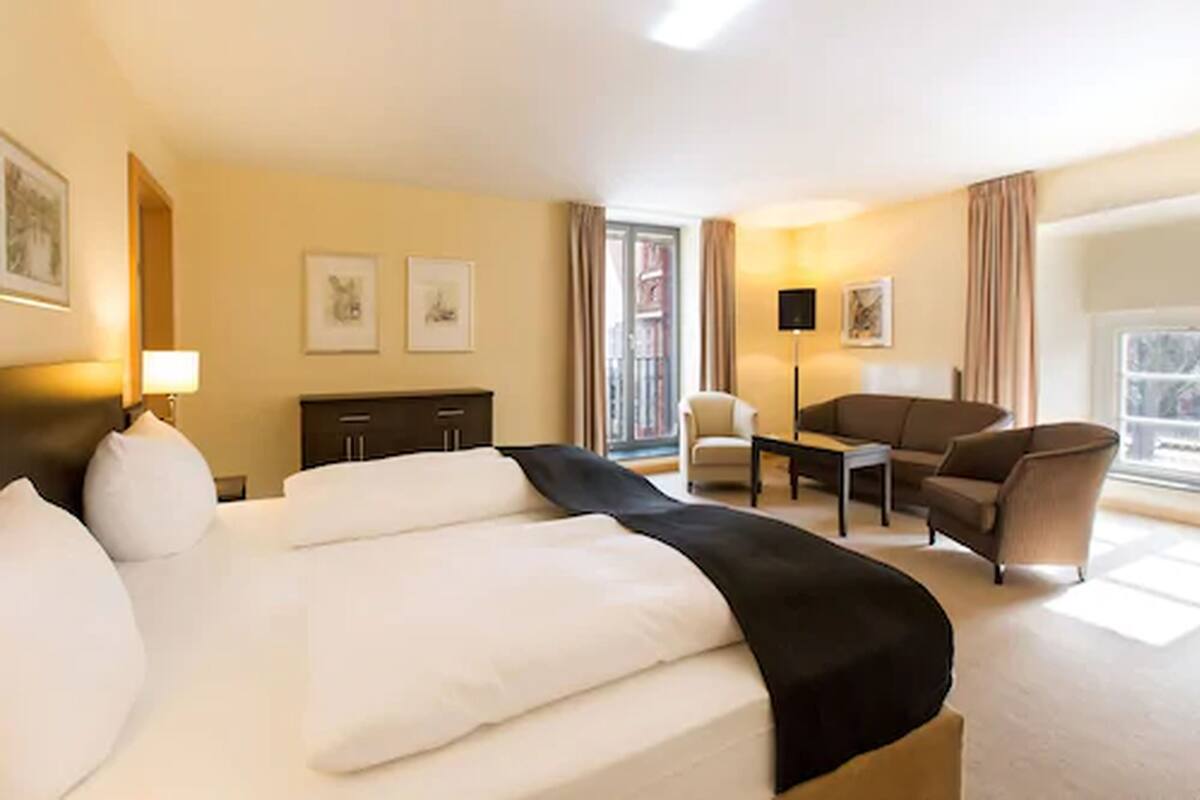 Une chambre d’hôtel avec un grand lit, un bureau, une chaise et un canapé. Il y a deux tableaux encadrés au mur, une lampe et une fenêtre avec des rideaux.