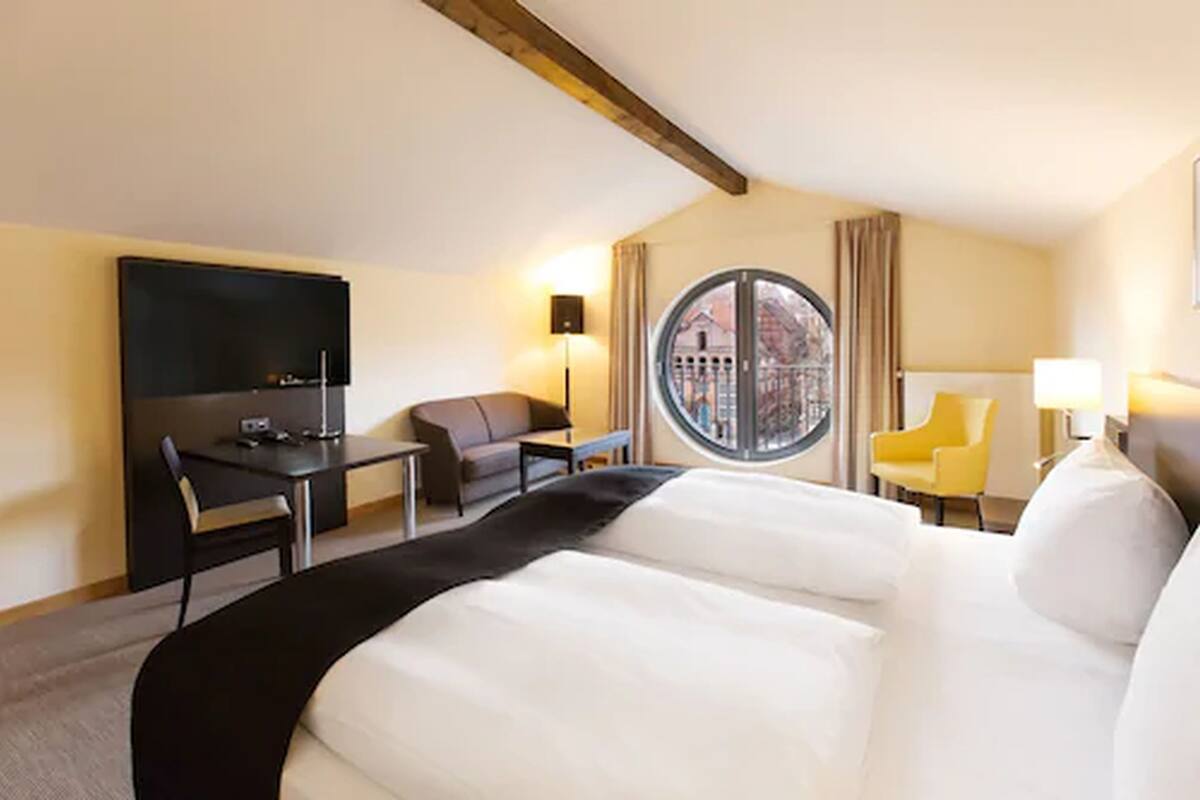 Une chambre d’hôtel avec un grand lit, un bureau, une chaise, une télévision et une fenêtre donnant sur des immeubles.
