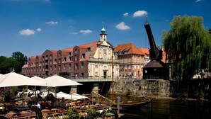A riverside café with outdoor seating, a historic building with a tower, and a bridge in the background. - DORMERO Hotel Altes Kaufhaus Lüneburg (Lueneburg)