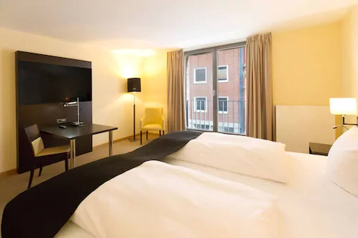 Une chambre d’hôtel avec un grand lit, un bureau, une chaise, une télévision et une fenêtre avec des rideaux.