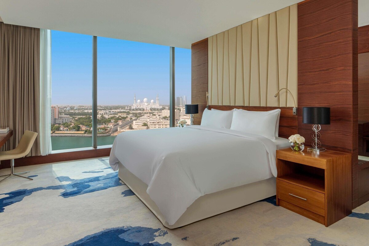 A hotel room with a large bed, a wooden nightstand, a desk, and a chair. The room has a large window with a view of a cityscape and a body of water.