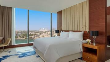 A hotel room with a large bed, a wooden nightstand, a desk, and a chair. The room has a large window with a view of a cityscape and a body of water.