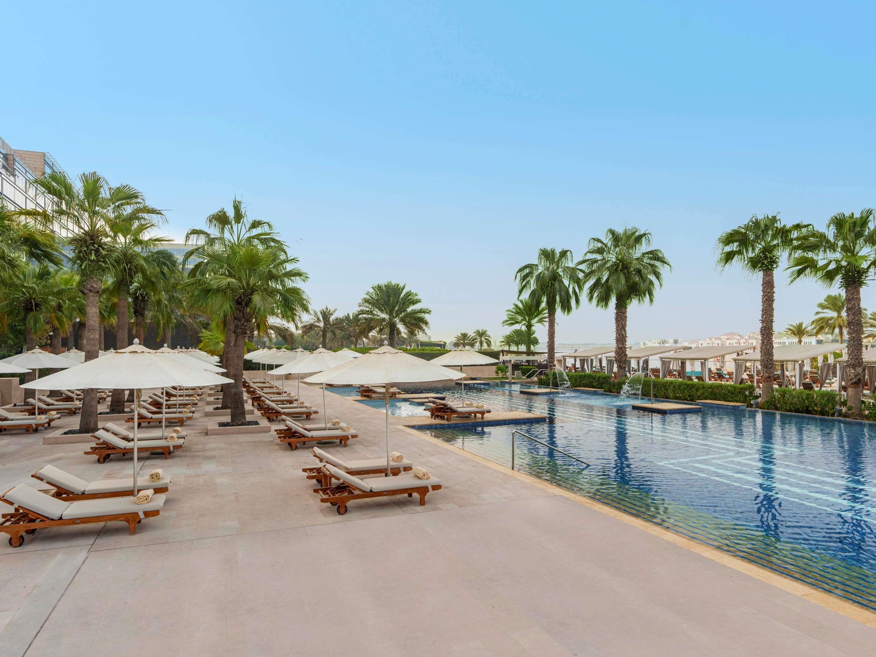 A poolside area with lounge chairs and umbrellas, palm trees, and a clear sky.