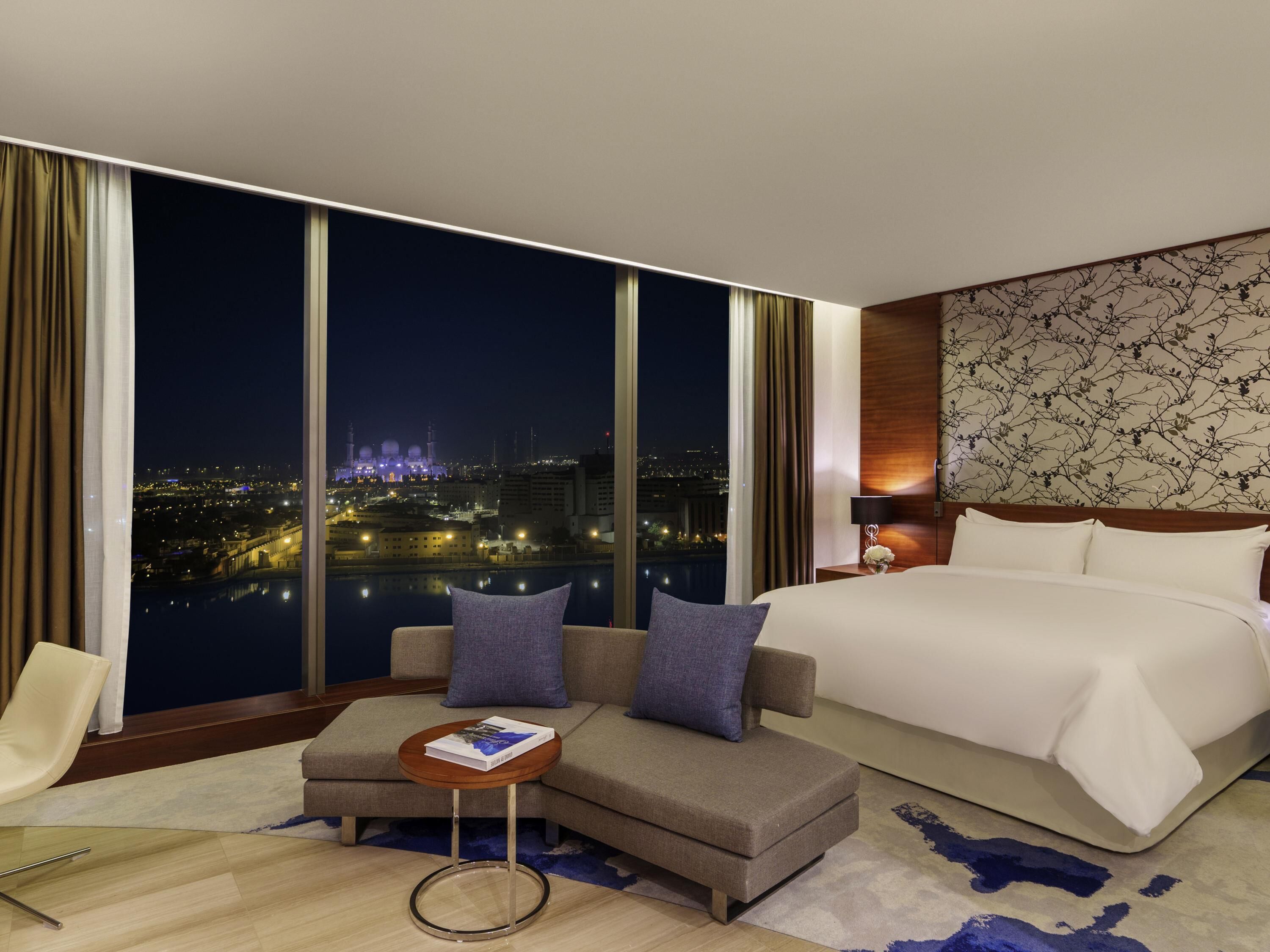 A modern hotel room with a large bed, a sofa, a small table, and a view of a cityscape at night.