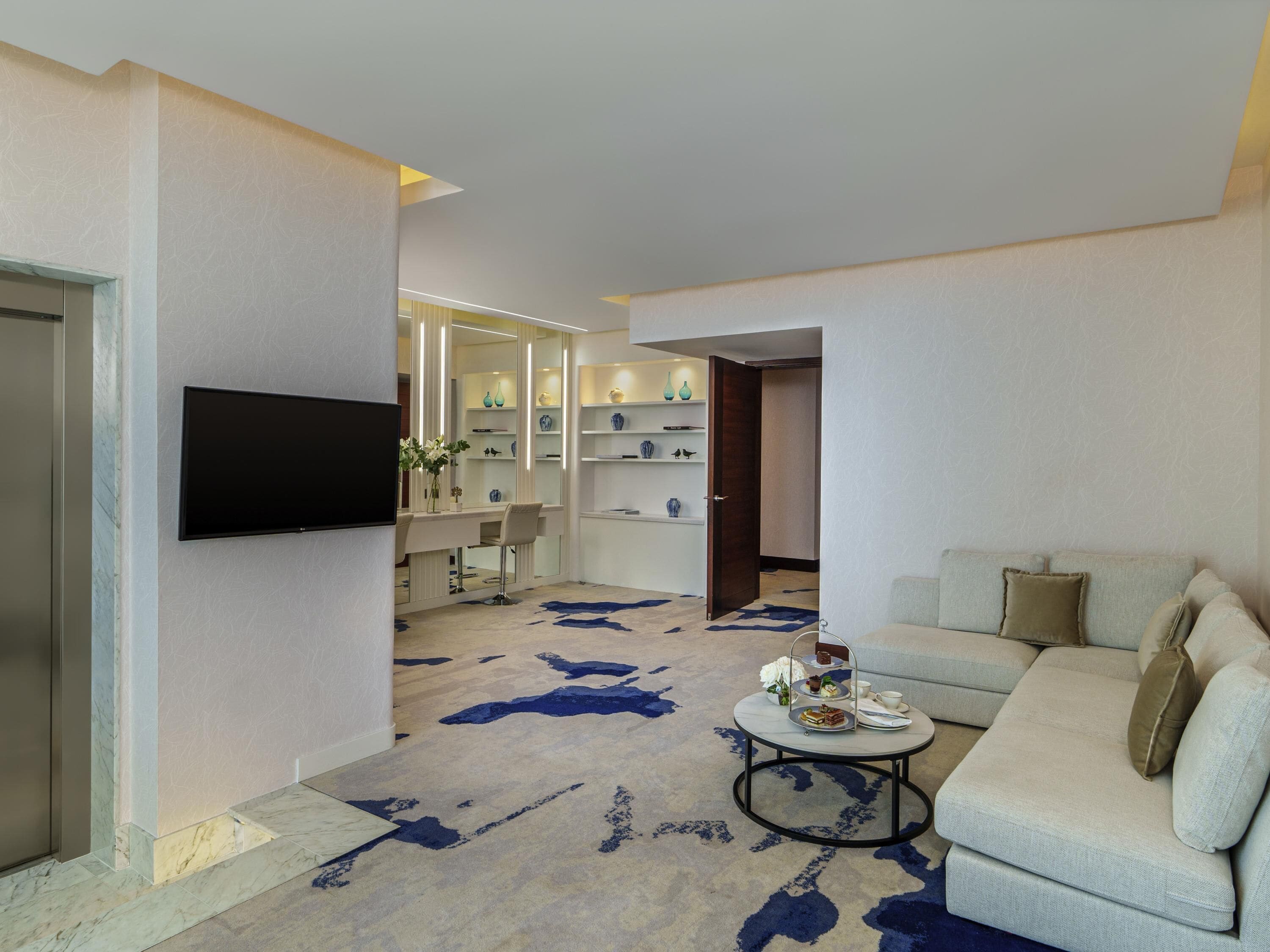 A modern living room with a sofa, a coffee table, a TV mounted on the wall, and a hallway leading to a dining area with a mirrored wall and shelves.