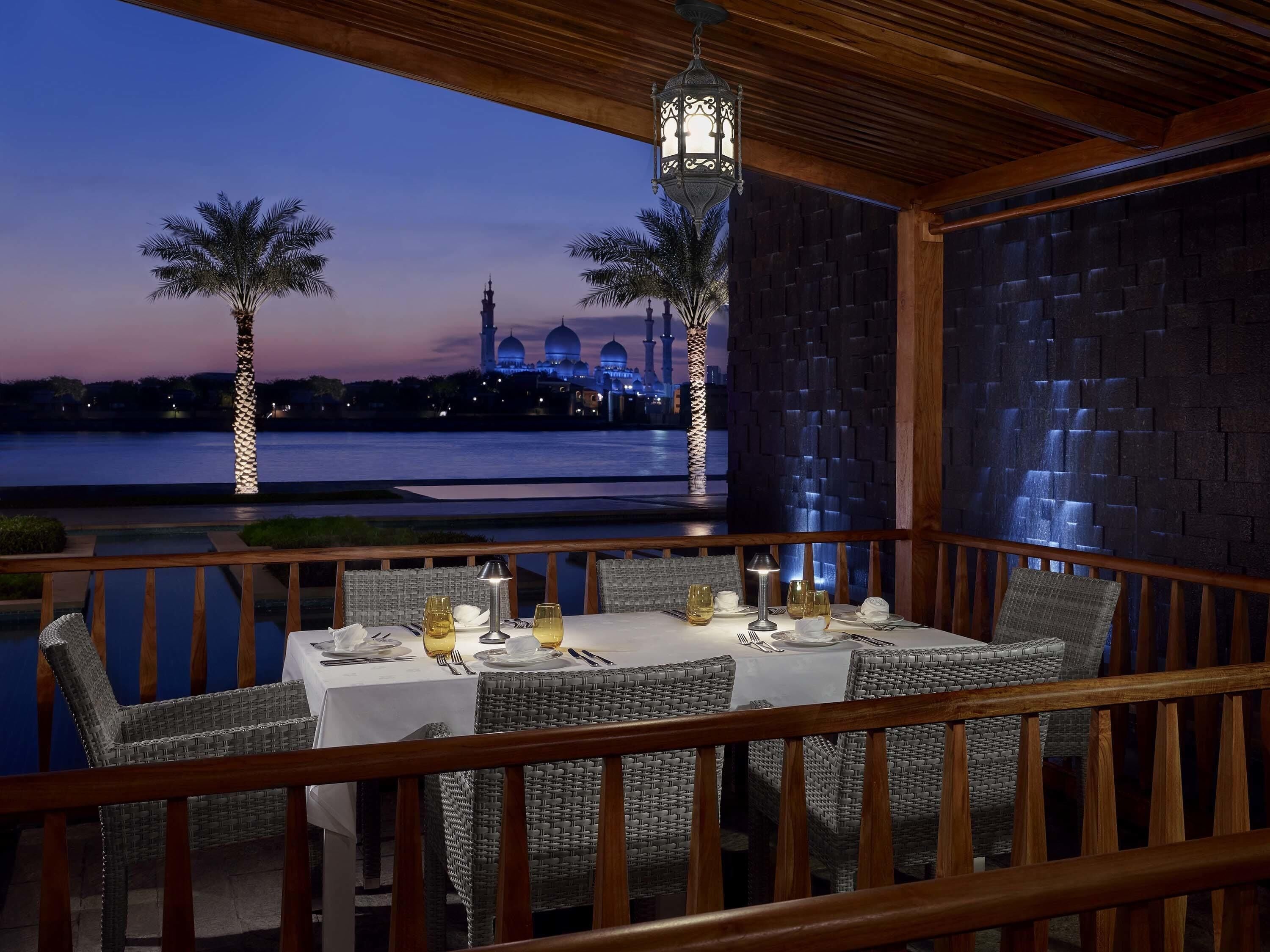 A dining area with wicker chairs, a table set for two, and a view of a mosque with domes and minarets.