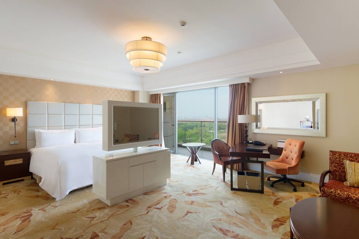 A hotel room with a bed, a TV, a desk with a chair, a large window, and a view of the city.