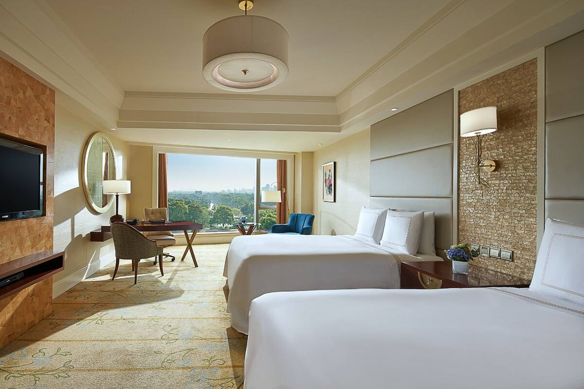 A hotel room with two beds, a desk with a chair, a television, and a large window offering a city view.