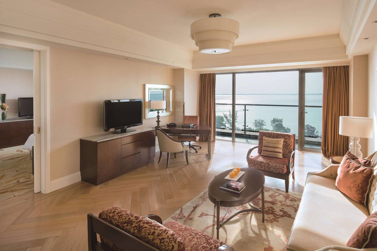 A spacious living room with a large window offering a view of the sea, a flat-screen TV, a dining area, and a sofa with a patterned throw.